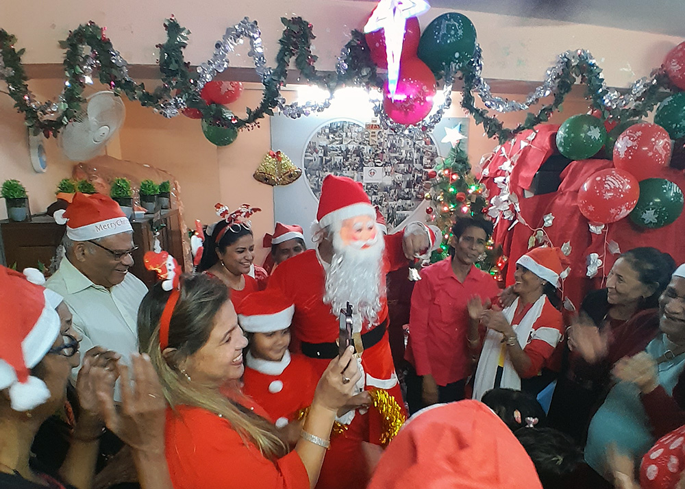 A group of people wearing Santa hats and one wearing a Santa mask smiling and clapping in a room adorned with Christmas decorations