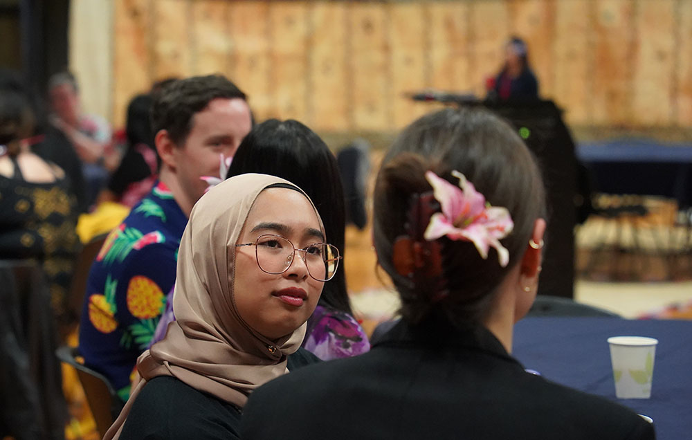 A woman wearing a headscarf listening to a woman with a flower in her hair in a hall with tapa cloth on the walls