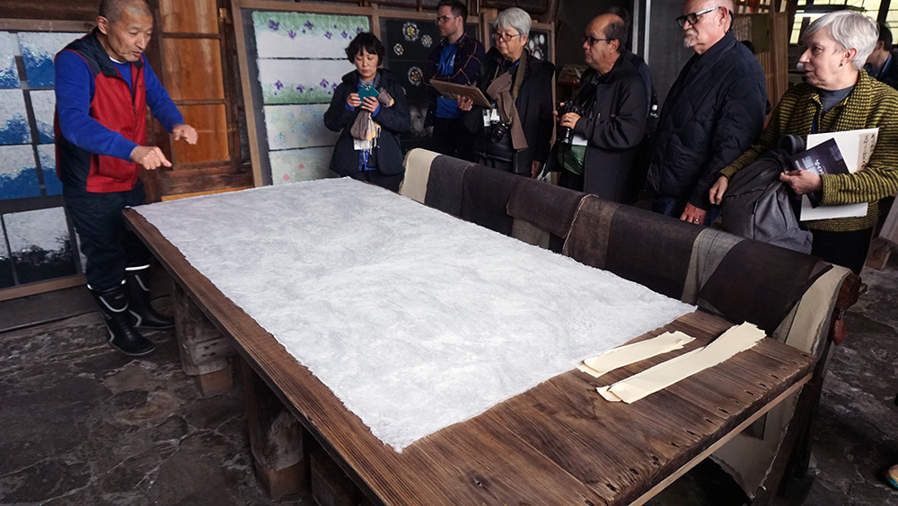 A group of people looking at a sheet of white textured paper laid out on a table  