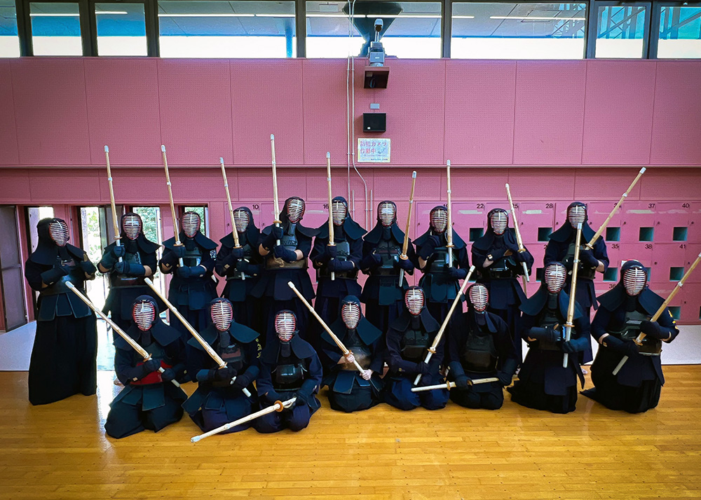 The team of 18 girls and staff dressed in kendo protective clothing and holding bamboo kendo swords