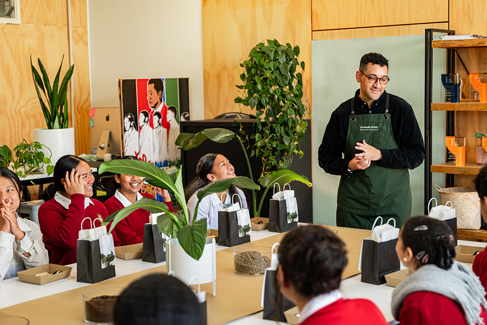 Joshua standing at the front of a classroom of young students with plants on their desks