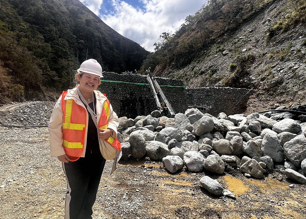 Krychelle wearing hi-vis jacket and hardhat in front of a small hydropower plant under construction