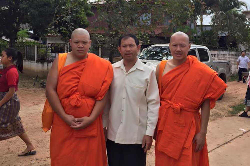 Three men stand in Laos streets, two in traditional orange robes