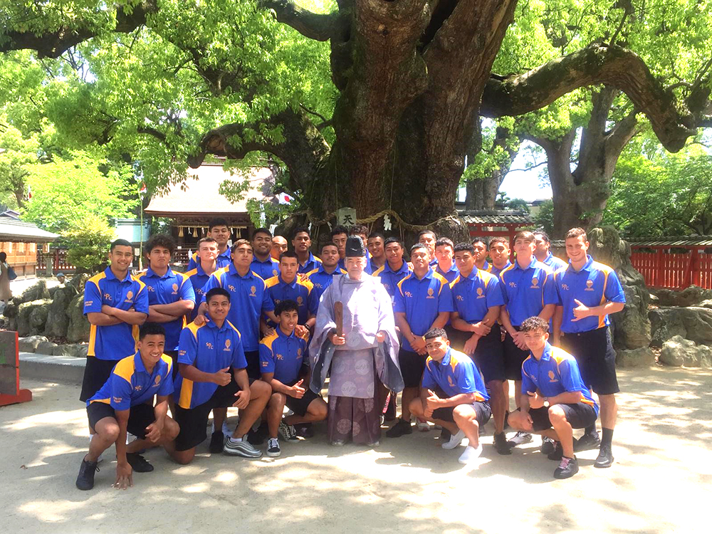 The team posing beneath a large tree with a Shinto priest