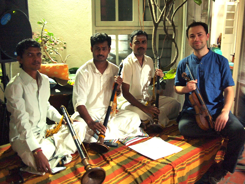 Tristan holding his violin while sitting with a group of three Indian musicians holding their instruments