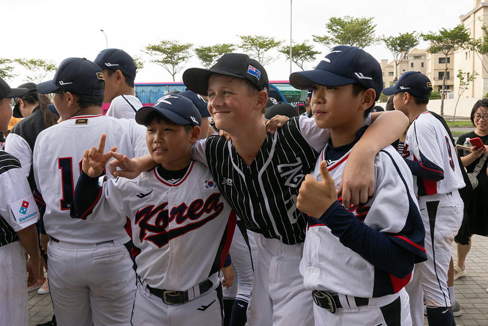 A young NZ baseball players with his arms around two Korean players