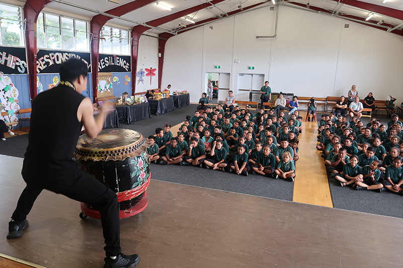 A drummer on stage drumming in front of a hall full of students