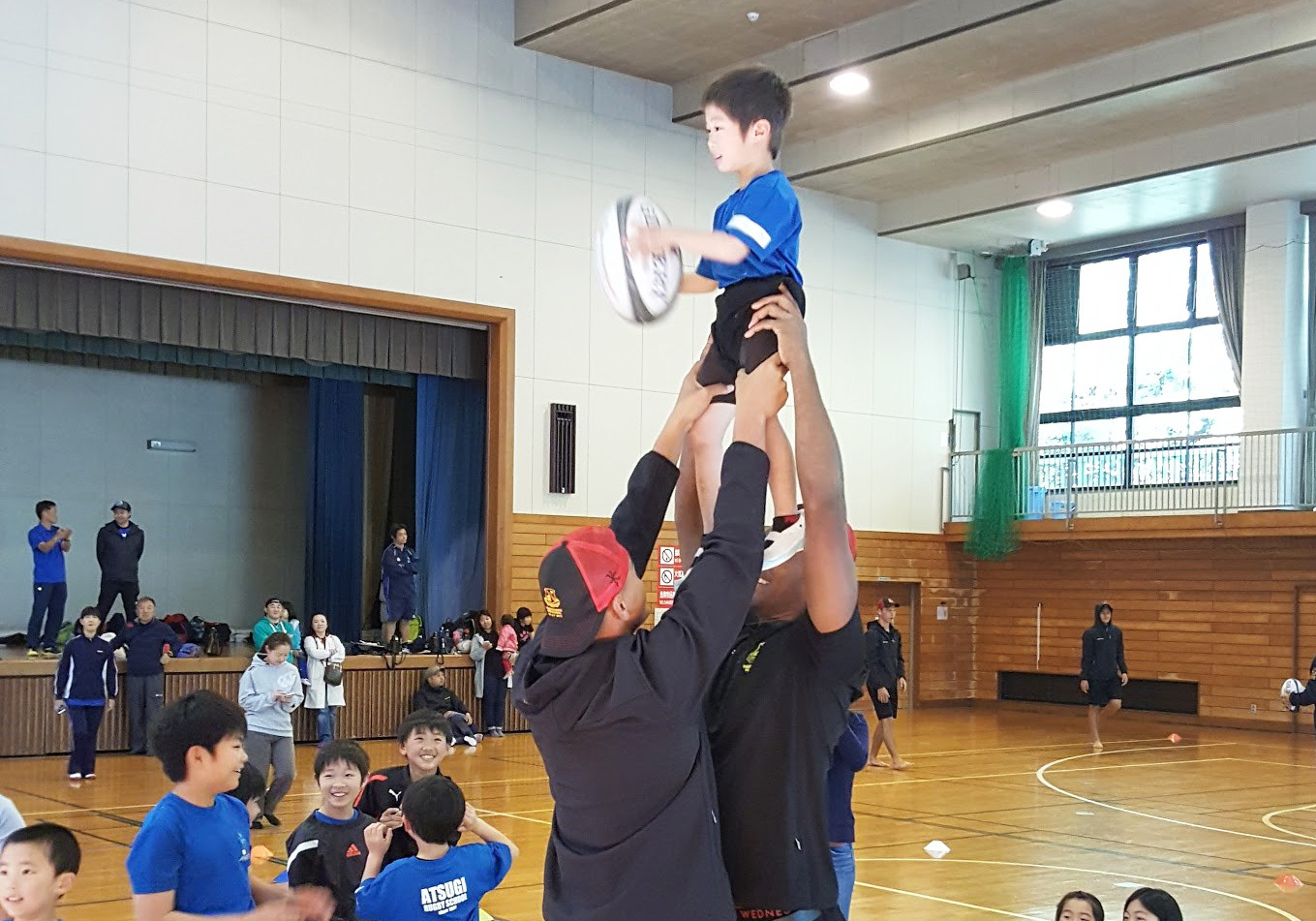 Two Hamilton Boys Rugby players lift a young boy into the air to catch a rugby ball