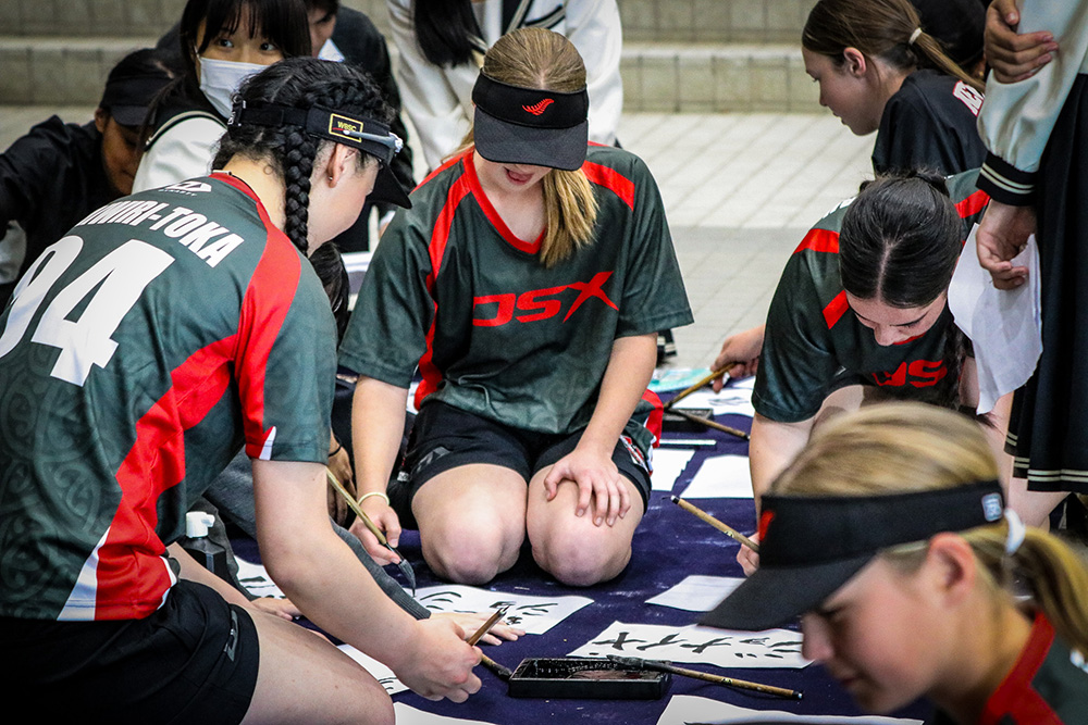Girls in their softball uniforms sitting on the ground practicing Japanese calligraphy