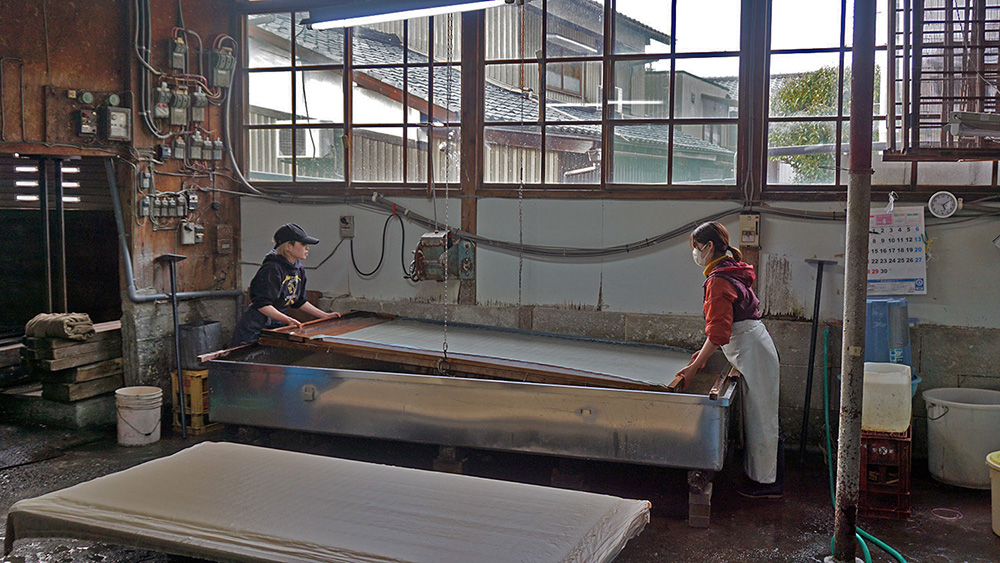 Two women in a factory lifting a very large sheet of paper out of a tub where it is being prepared 