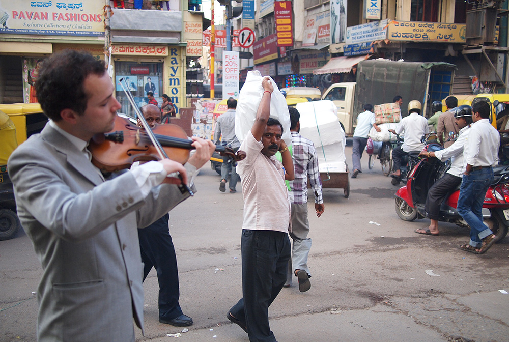 A man carrying a sack on his sholder watches Tristan play the violin on a street in Bangalore