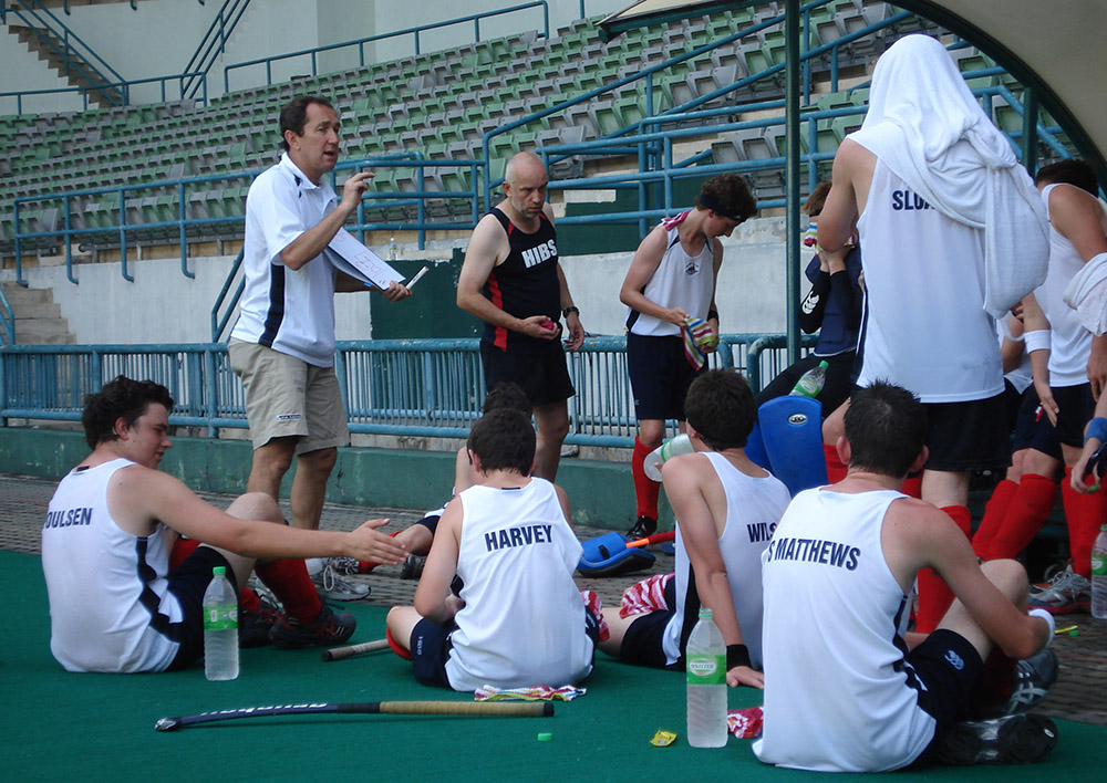 Peter talking to a group of young hockey player who are sitting on turf with their hockey sticks