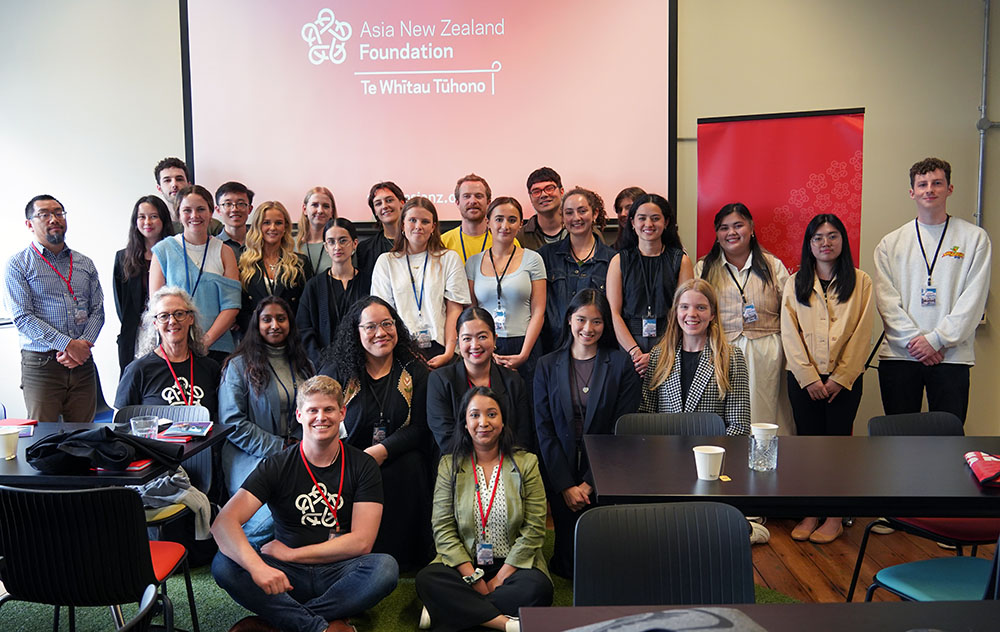 A group shot of about 25 interns and handful of staff posing in front of an Asia New Zealand Foundation banner projected onto a screen