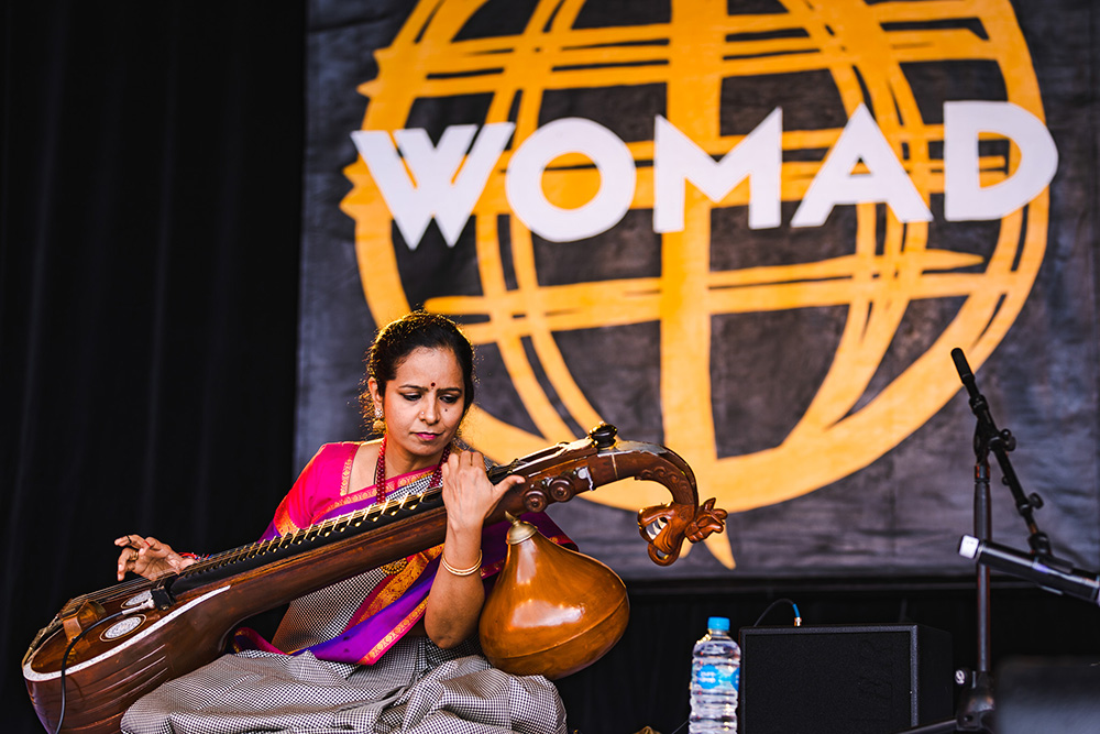A woman playing a sitar on stage with the WOMAD banner behind her