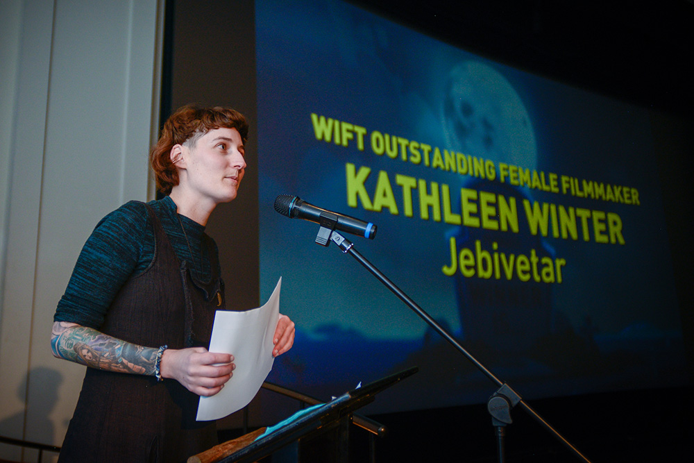 Kathleen standing at a microphone with her name displayed on a screen behind her
