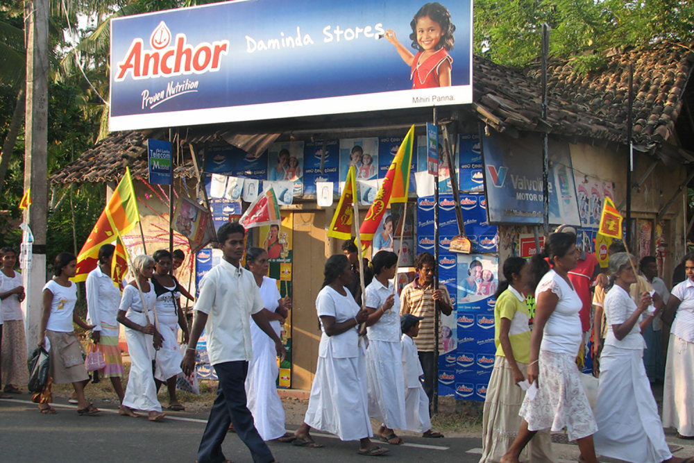 Group of young people walk down the street past an Anchor milk sign with flags