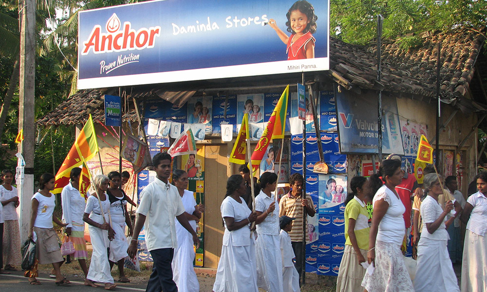 People dressed in white and carrying Sri Lankan flags walk past a sign for Ankor butter