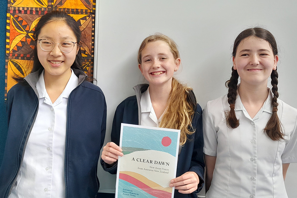 Three students smiling with the middle stutent holding a copy of the anthology of Asian New Zealand poetry A Clear Dawn