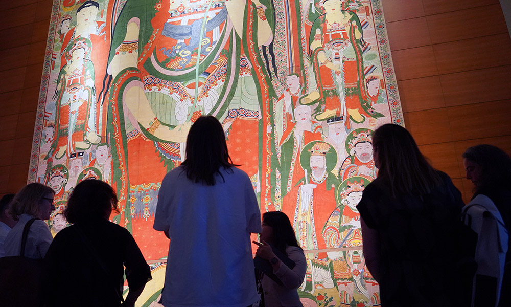 A group of five people looking at a large traditional Korean wall painting of figures in traditional dress