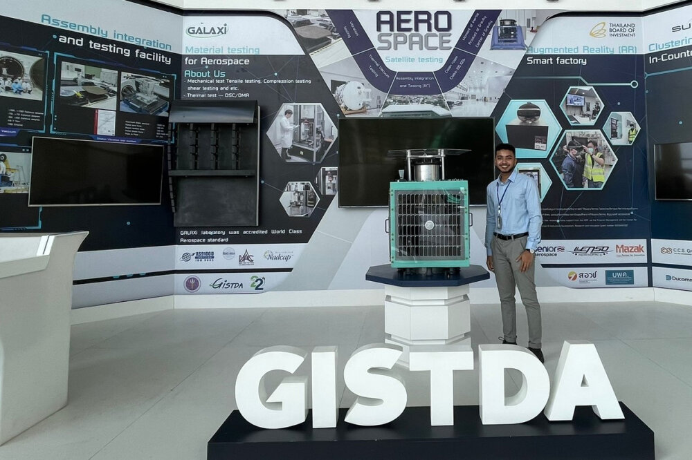 Waikato University student Siveshearn Jaynessh standing in a room with the GISTDA sign in front of large infographics displaying the work they do