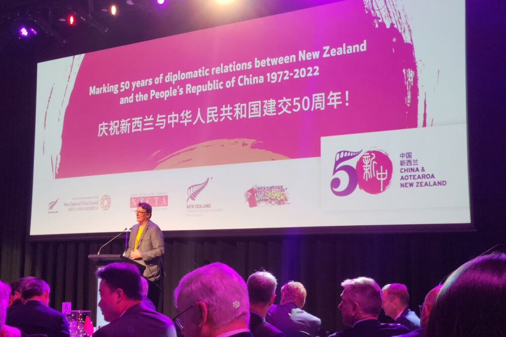 A man standing on a stage with a large pink sign behind him marking 50 years of diplomatic relations between New Zealand and China
