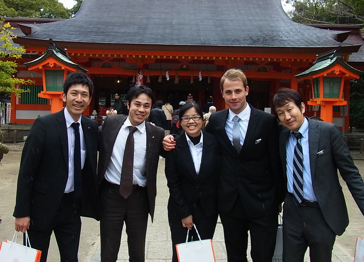 A group of five people wearing suits standing arm in arm in front of a Japanese temple