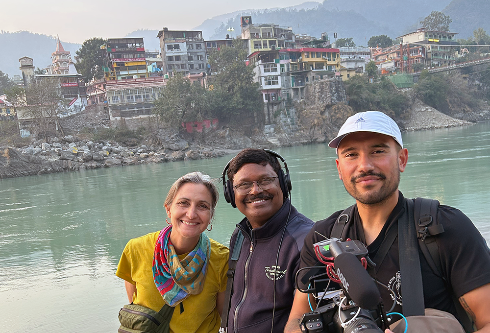 Three people holding camera gear with colourful buildings visible behind them on the other side of a large river