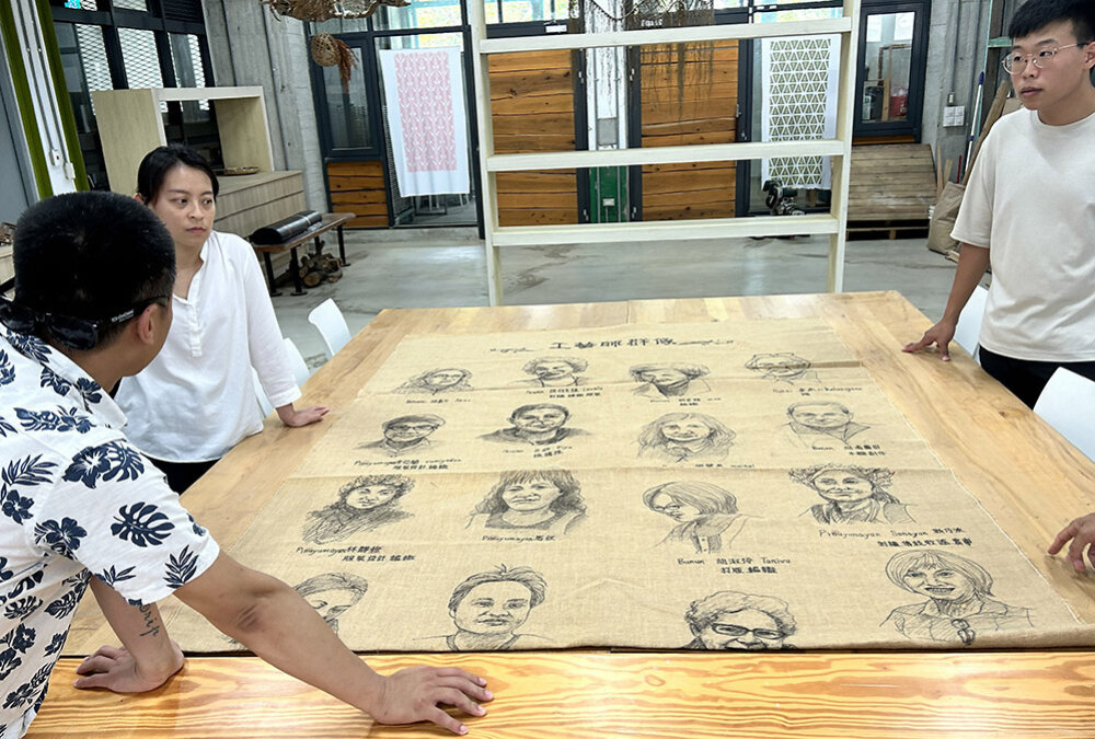 Three people standing around a table on which there is a large artwork depicting the faces of people