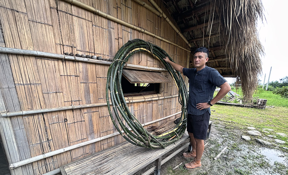 A man standing beside a thatched house holding lengths of bamboo bound together into a circle
