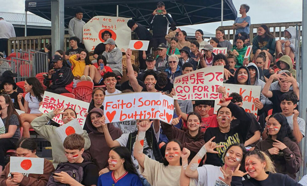 Students sitting in a stand waving and holding signs supporting the Japanese softball team