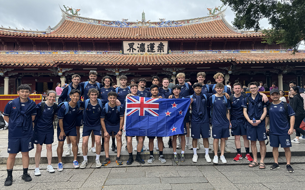 The full team posing for a photo in their uniforms and holding a New Zealand flag in front of the Kaiyuan Temple 