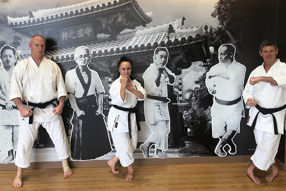 Three people in karategi (karate uniforms) posing in front of life-sized images of famous karate exponents