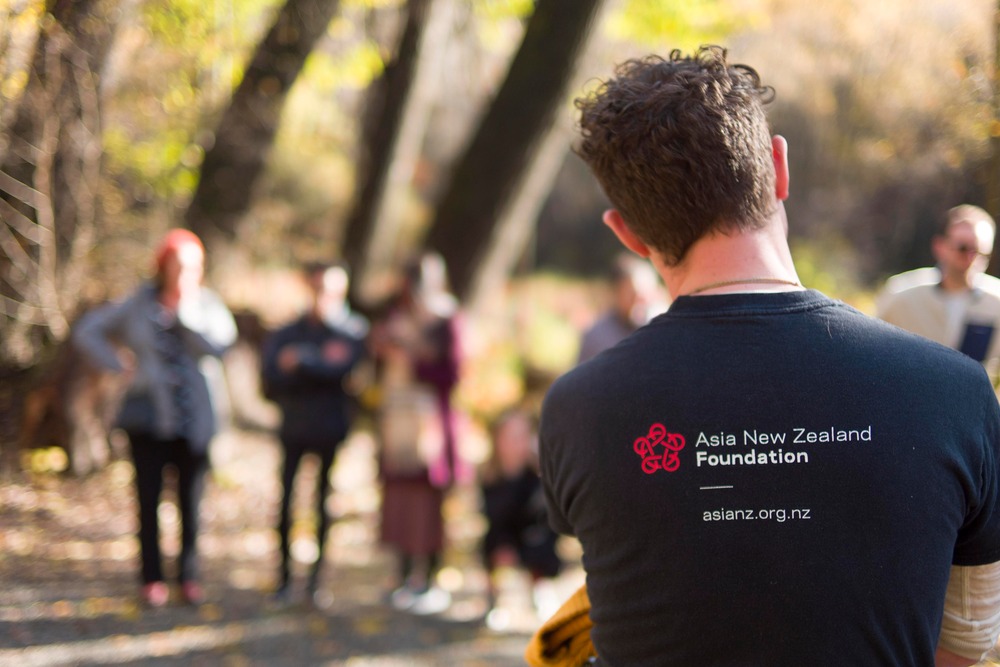A man wearing an Asia New Zealand Foundation t-shirt with Leadership Network members out of focus in the background