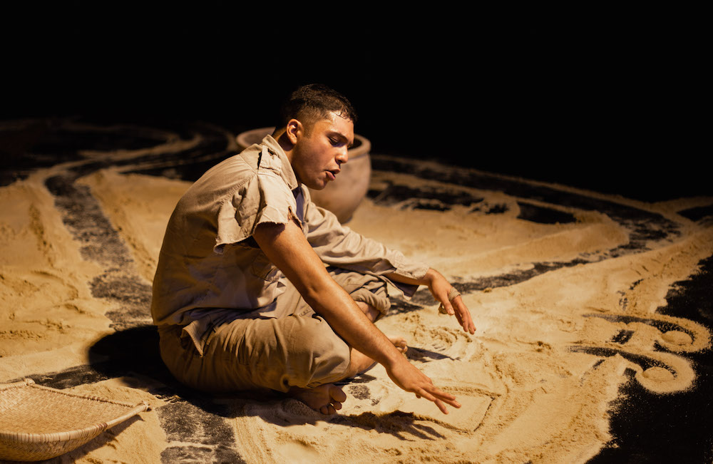 A man sitting on a stage floor making patterns in sand