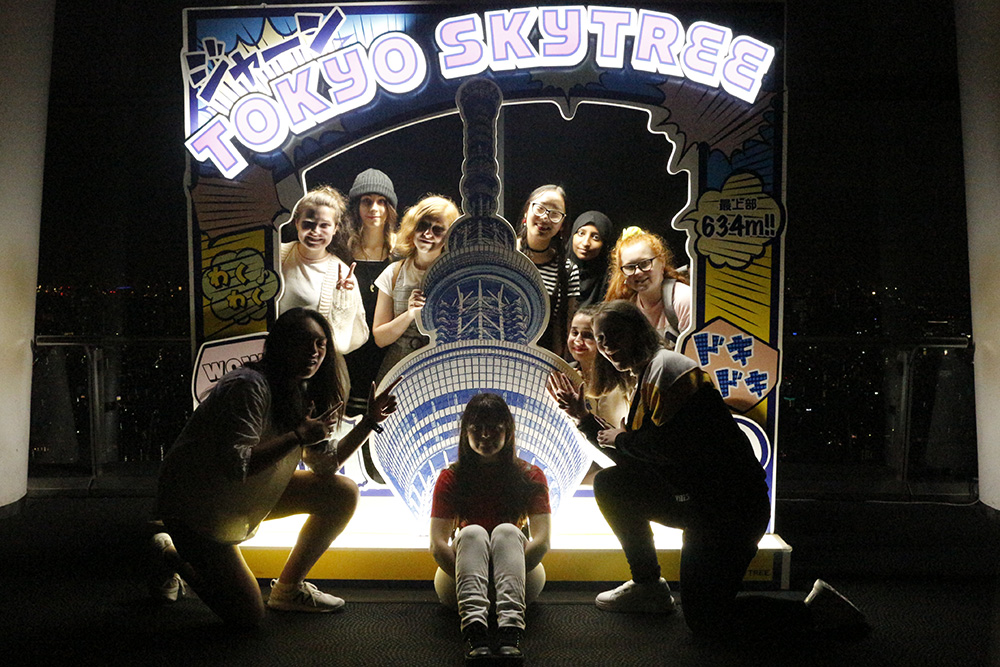 The Otago Girls High School students standing and sitting in front of a lit up sign for the Tokyo Skytree