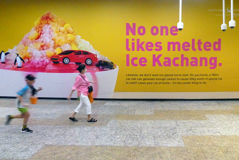A child and a woman walking past a billboard promoting public transport use that reads 'No one likes melting ice kachang'