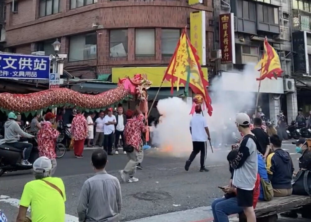 People watching a team performing a dragon dance on a city street, accompanied by the flash of fireworks