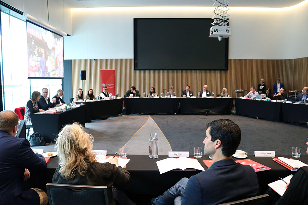 A large conference room with conference attendees sitting at tables arranged in a circle