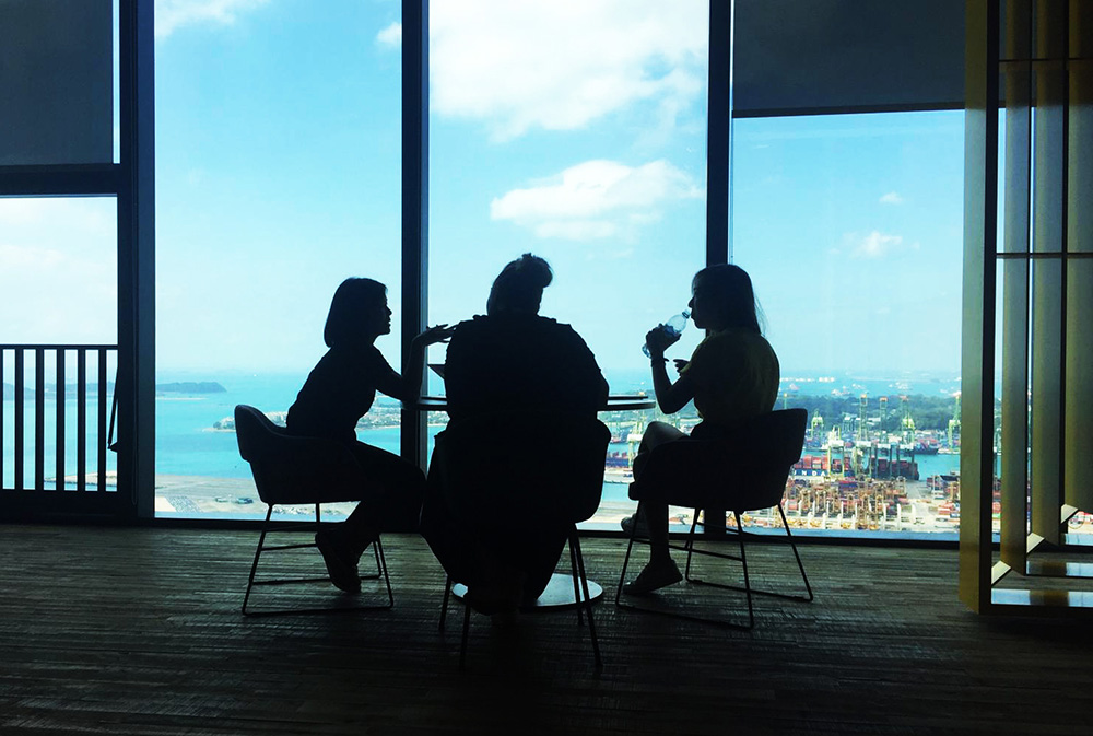 A silhouette image of three people sitting at a table in front of a big window overlooking a harbour