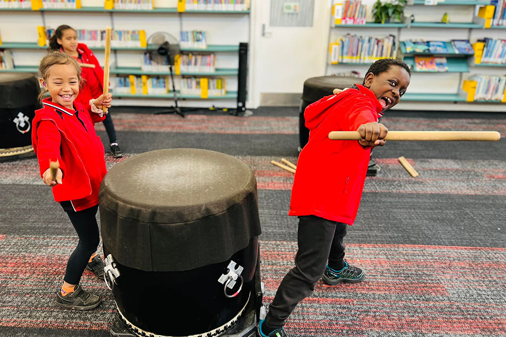 Two students standing beside a drum holding drum sticks and grinning