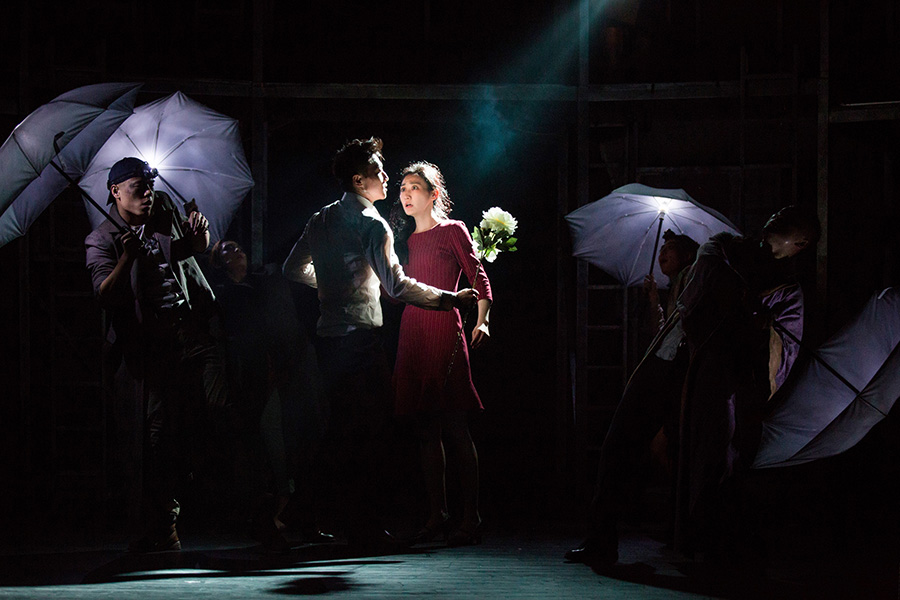 Two performers on stage under a spotlight while supporting actors stand with umbrellas around them
