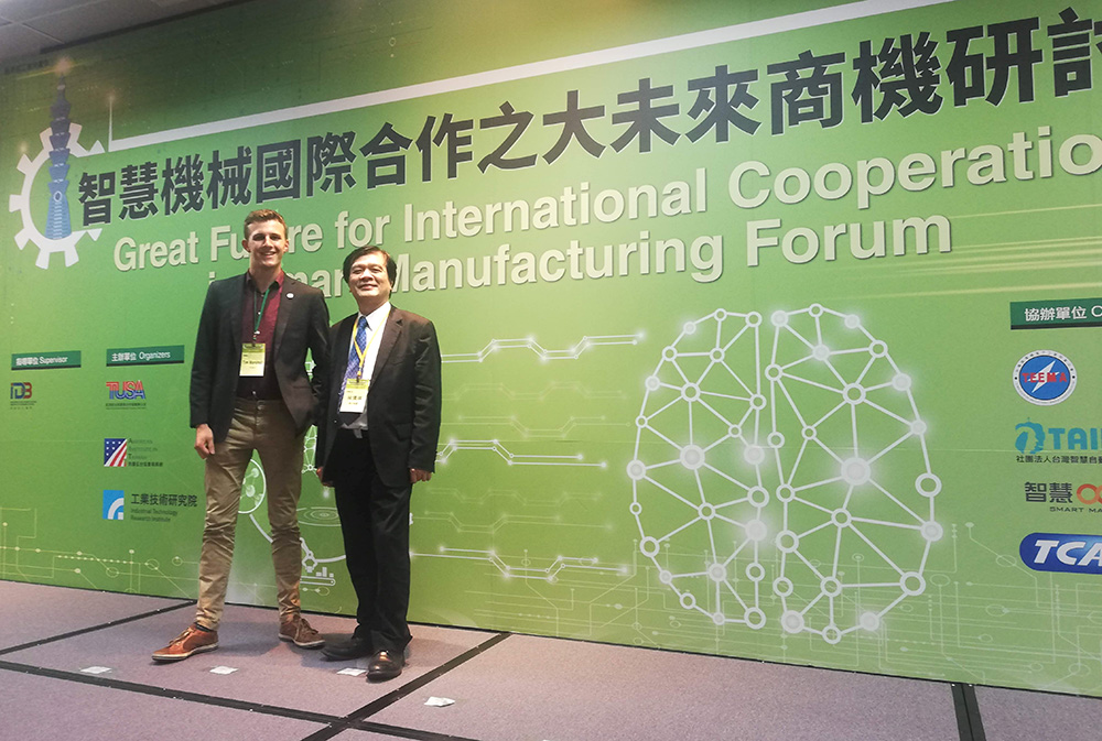 Tim standing with a colleague in front of a large sign promoting a business forum