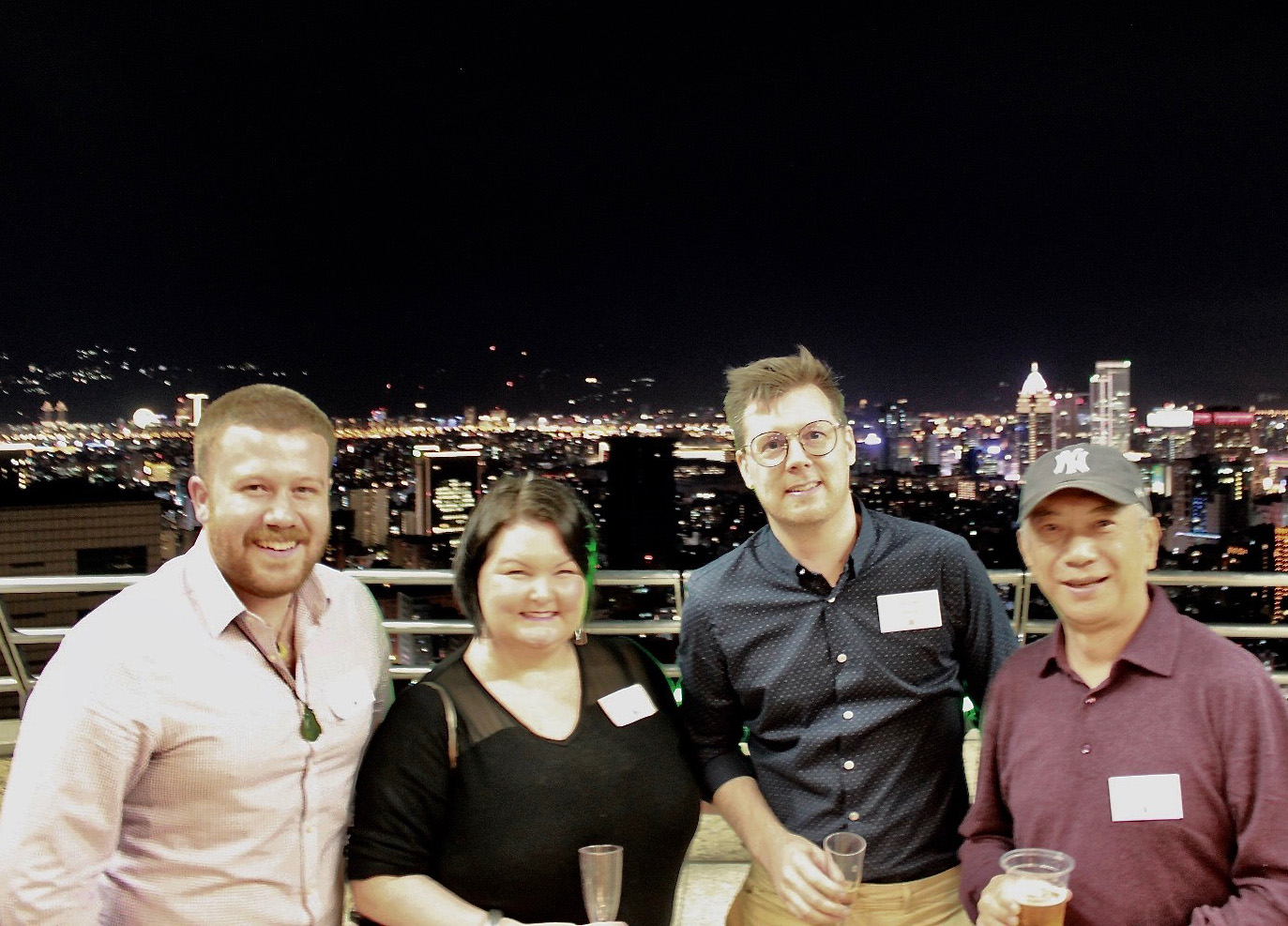 Ben standing oon a balcony at night with three colleagues with a city skyline at night in the background