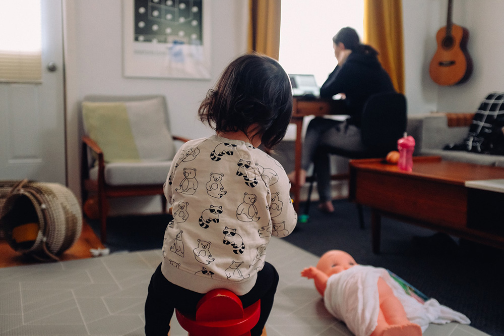 A toddler sitting on a stool with a woman working on a laptop in the background