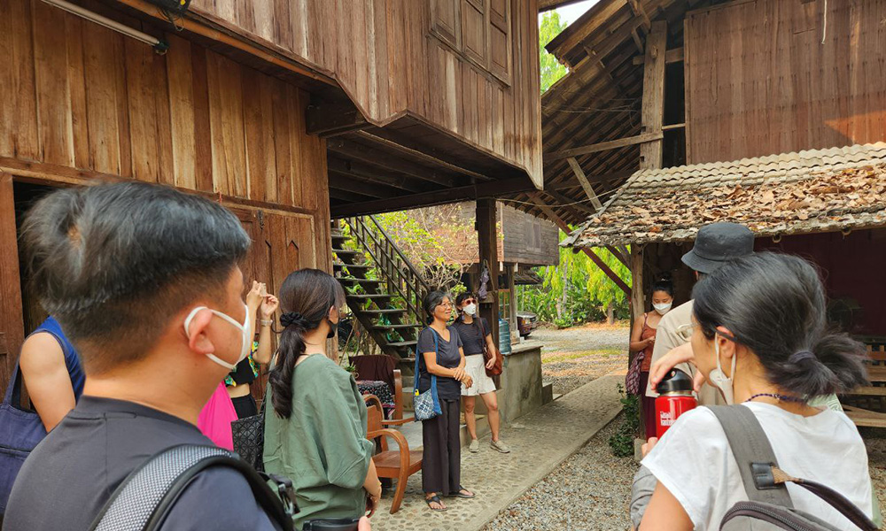 A woman speaking to a group beside traditional-style Thai houses