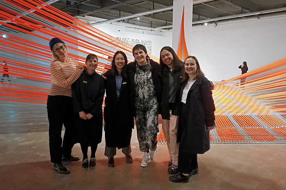 Five women (curators) standing in front of an installation of strips of orange tape