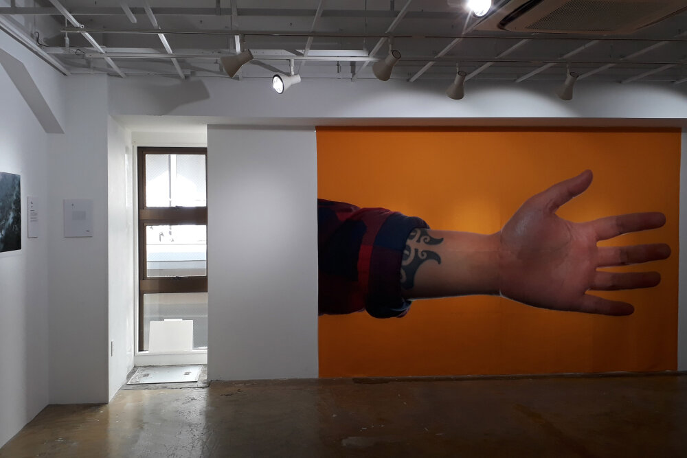 An image of a gallery space with a large photo of a personas hand and wrist taking up one wall