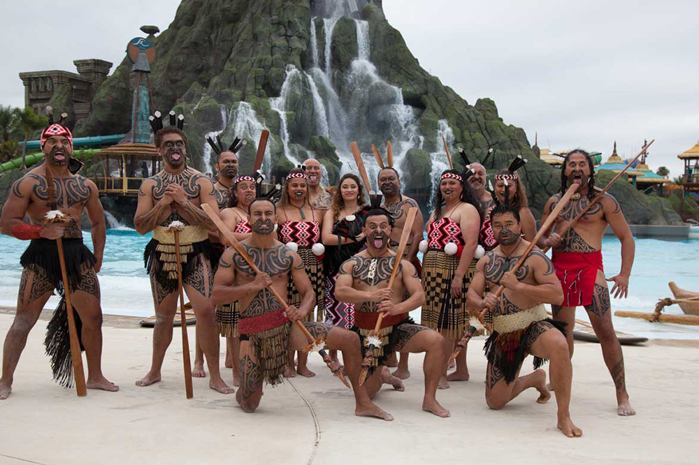 A group in traditional Maori dress in front of a fun park mountain and waterfall